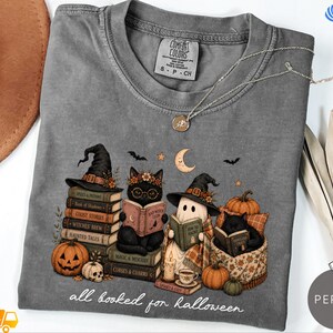 A gray t-shirt with a Halloween-themed graphic design featuring cats dressed as witches and wizards, along with various Halloween-related items such as pumpkins, bats, and a crescent moon.