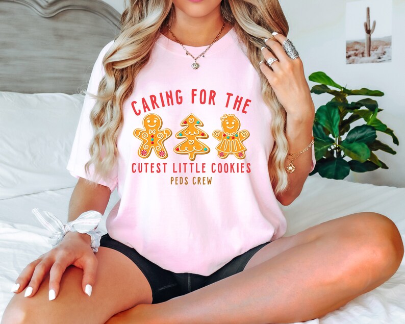 以下が含まれることがあります： 「Caring for the Cutest Little Cookies Peds Crew」というテキストと、ジンジャーブレッドマン、ジンジャーブレッドツリー、ジンジャーブレッドガールが描かれたピンクのTシャツです。