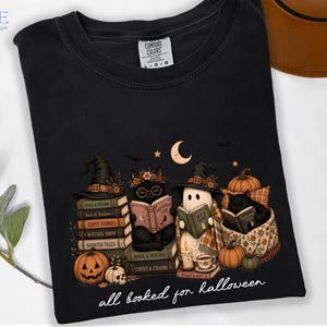 A black t-shirt with a Halloween-themed graphic design depicting a witch, a ghost, and a cat reading books, surrounded by pumpkins, bats, and a crescent moon.