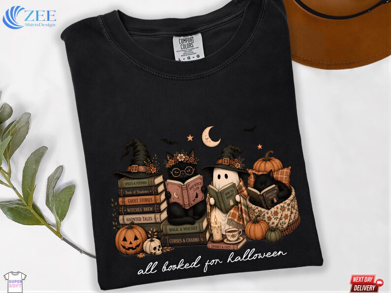 A black t-shirt with a Halloween-themed graphic design depicting a witch, a ghost, and a cat reading books, surrounded by pumpkins, bats, and a crescent moon.