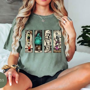 May include: A moss green short-sleeve t-shirt featuring a graphic print of four vertical panels. The panels display characters from The Nightmare Before Christmas, including Sally, Oogie Boogie, Jack Skellington, and Lock, Shock, and Barrel.