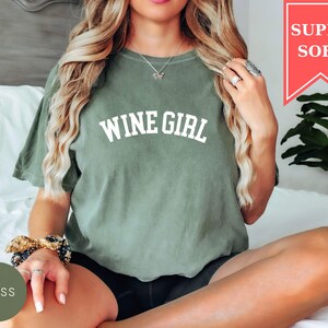 Pode incluir: Uma camiseta verde musgo com o texto "WINE GIRL" em letras brancas. A camisa tem um ajuste folgado e &eacute; feita de um material macio. Uma faixa vermelha no canto superior direito diz "SUPER SOFT".