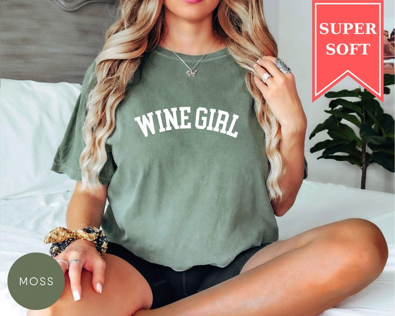 Pode incluir: Uma camiseta verde musgo com o texto "WINE GIRL" em letras brancas. A camisa tem um ajuste folgado e &eacute; feita de um material macio. Uma faixa vermelha no canto superior direito diz "SUPER SOFT".