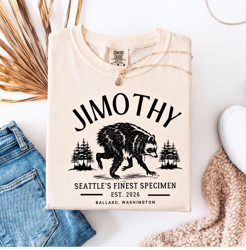 A beige t-shirt with a graphic design featuring a bear and pine trees, along with text that reads "Jimothy, Seattle's Finest Specimen, EST. 2026, Ballard, Washington".