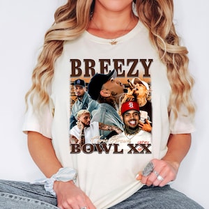 May include: Cream-colored t-shirt featuring a graphic of Chris Brown with the text "BREEZY" above and "BOWL XX" below. The design includes multiple images of the artist. The shirt is worn by a person.
