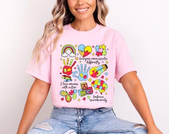 May include: Pink t-shirt with a colorful graphic design featuring puzzle pieces, a rainbow, handprints, a lightbulb, and the text "Everyone communicates differently", "I love someone with autism", "ABC", and "Embrace neurodiversity".