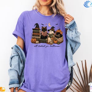 A woman wearing a purple t-shirt with a Halloween-themed graphic of cats and a book, along with the text "all baked for halloween".