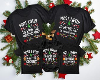May include: Set of four black t-shirts with Christmas-themed text and graphics. The shirts read "Most Likely to Sing the Christmas Songs", "Most Likely to Watch All the Football Games", "Most Likely to Eat the Cookies", and "Most Likely to Wake Up First".