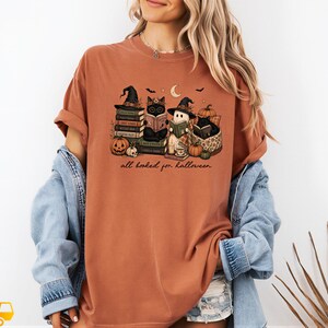 A woman wearing a t-shirt with a Halloween-themed graphic design, including books, pumpkins, bats, and a cat.