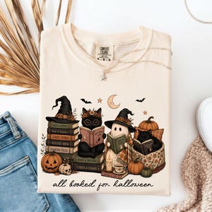 A white t-shirt with a Halloween-themed graphic design, featuring three cats dressed as witches and a ghost, surrounded by books, pumpkins, and other Halloween-related items.