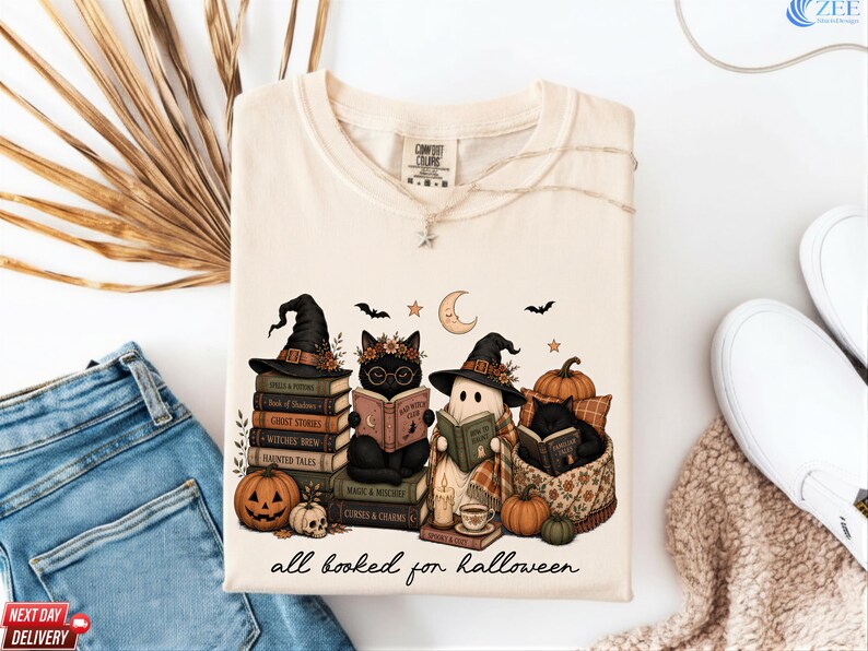 A white t-shirt with a Halloween-themed graphic design, featuring three cats dressed as witches and a ghost, surrounded by books, pumpkins, and other Halloween-related items.