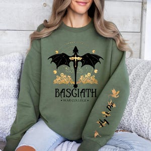 May include: A green sweatshirt with a black dragon graphic and gold accents. The text "BASGIATH - WAR COLLEGE -" is printed on the front of the sweatshirt. The sleeve has a gold dragon graphic and the text "Fly At Die".