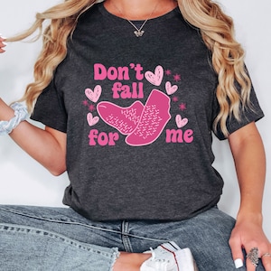 Op de afbeelding: Een donkergrijs T-shirt met de tekst "Don't fall for me" in roze letters. De tekst wordt omringd door roze harten en sterren.