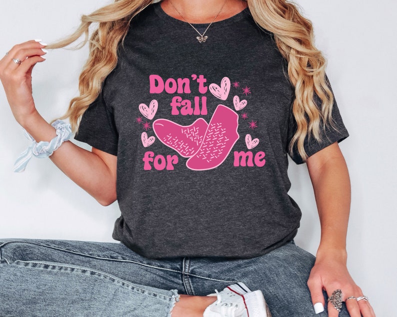 Op de afbeelding: Een donkergrijs T-shirt met de tekst "Don't fall for me" in roze letters. De tekst wordt omringd door roze harten en sterren.