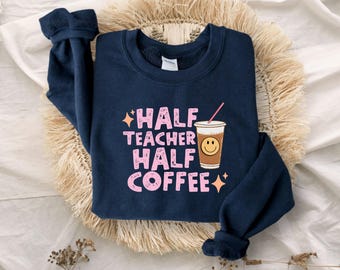 Op de afbeelding: Een marineblauw sweatshirt met lange mouwen en de roze, verweerde tekst "HALF TEACHER HALF COFFEE", vergezeld van een afbeelding van een koffiebeker met een smiley en een roze rietje, geaccentueerd door kleine oranje vierpuntige sterren.