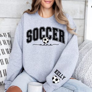 May include: Gray sweatshirt with the text "SOCCER mama" and a soccer ball graphic. The left sleeve has the text "Kelly" and a soccer ball graphic.