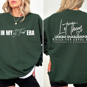 May include: A dark green sweatshirt with white text on the front and back. The front text reads "In My Let Them Era". The back text reads "Let Them Keep Talking While You Level Up".