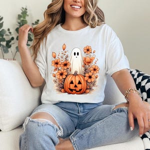 May include: White t-shirt featuring a Halloween-themed graphic. The design includes a cartoon ghost, a jack-o'-lantern, and orange flowers. The shirt is made of a soft material and has short sleeves.