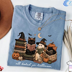 A blue t-shirt with a Halloween-themed graphic design, featuring cats dressed as witches and wizards sitting on books and surrounded by pumpkins, bats, and a crescent moon.