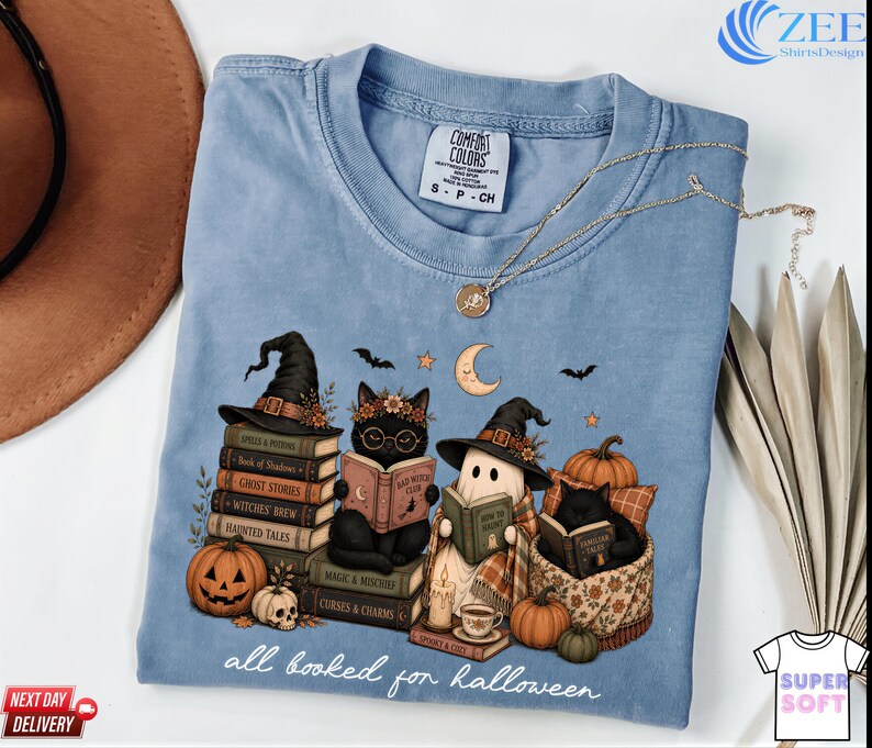 A blue t-shirt with a Halloween-themed graphic design, featuring cats dressed as witches and wizards sitting on books and surrounded by pumpkins, bats, and a crescent moon.