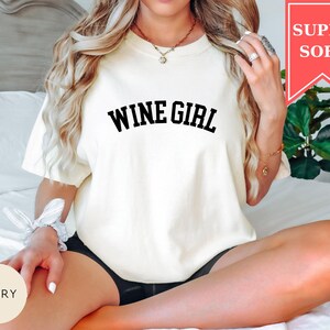 Pode incluir: Camiseta cor marfim com a frase "WINE GIRL" em letras pretas em negrito. A camiseta tem mangas curtas e um corte folgado. Uma faixa vermelha no canto superior direito diz "SUPER SOFT".