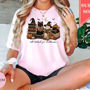 A woman wearing a pink t-shirt with a Halloween-themed graphic of cats and pumpkins, sitting on a bed with a potted plant in the background.