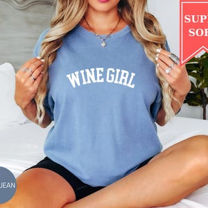 Pode incluir: Uma camiseta azul jeans com o texto "WINE GIRL" em letras brancas. A camisa tem mangas curtas e um ajuste relaxado. Uma faixa vermelha no canto superior direito diz "SUPER SOFT".