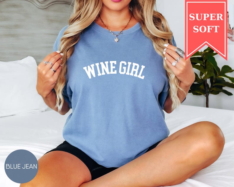 Pode incluir: Uma camiseta azul jeans com o texto "WINE GIRL" em letras brancas. A camisa tem mangas curtas e um ajuste relaxado. Uma faixa vermelha no canto superior direito diz "SUPER SOFT".