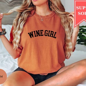 Pode incluir: Camiseta laranja com a frase "WINE GIRL" em preto. A camiseta tem um caimento folgado e &eacute; rotulada "SUPER SOFT" em uma faixa vermelha. A modelo est&aacute; usando shorts pretos e uma scrunchie floral.