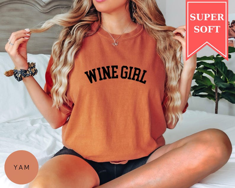 Pode incluir: Camiseta laranja com a frase "WINE GIRL" em preto. A camiseta tem um caimento folgado e &eacute; rotulada "SUPER SOFT" em uma faixa vermelha. A modelo est&aacute; usando shorts pretos e uma scrunchie floral.