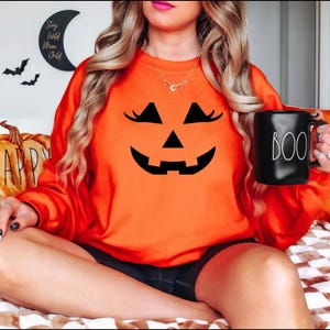 May include: A bright orange crewneck sweatshirt featuring a black jack-o'-lantern face graphic with eyelashes on the front. The sweatshirt is paired with a black ceramic mug that has the word BOO printed in white lettering on the side.