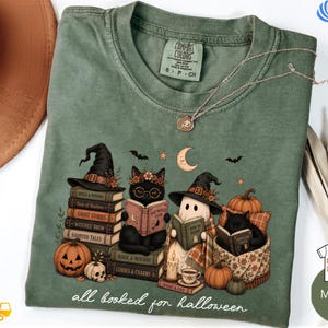 A green t-shirt with a Halloween-themed illustration of cats reading books, surrounded by pumpkins, bats, and other Halloween-related items.