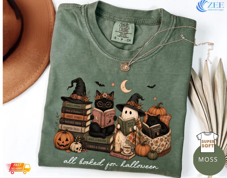 A green t-shirt with a Halloween-themed illustration of cats reading books, surrounded by pumpkins, bats, and other Halloween-related items.