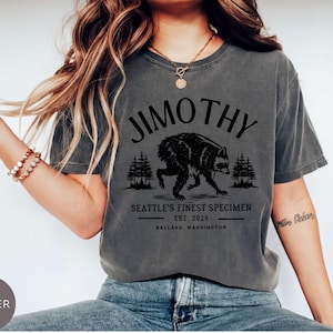 A woman wearing a gray t-shirt with a graphic design of a bear and text that says "Jimothy, Seattle's Finest Specimen, Est. 2026, Ballard, Washington".