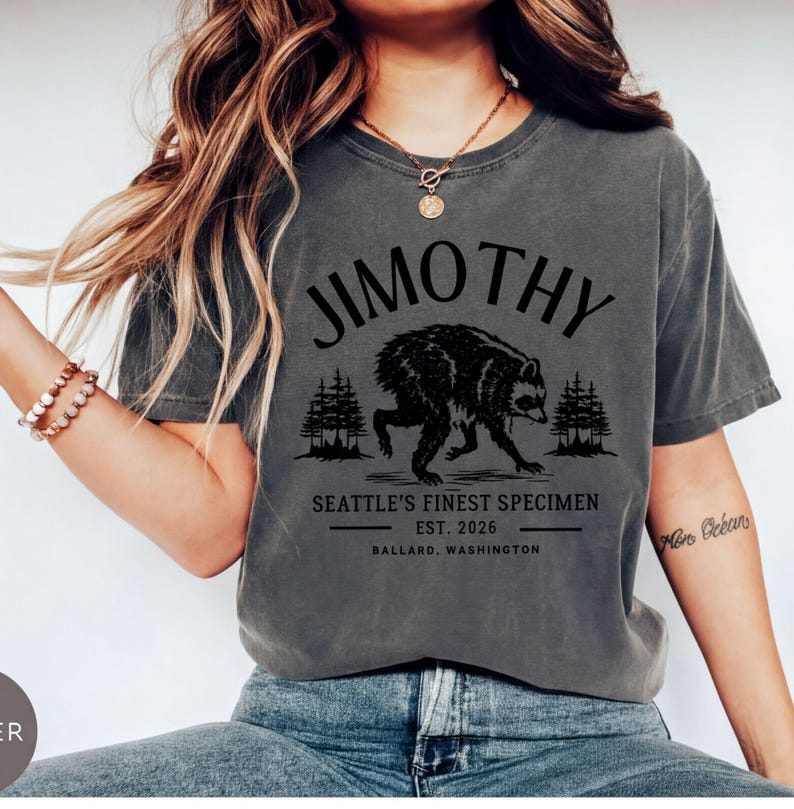 A woman wearing a gray t-shirt with a graphic design of a bear and text that says "Jimothy, Seattle's Finest Specimen, Est. 2026, Ballard, Washington".