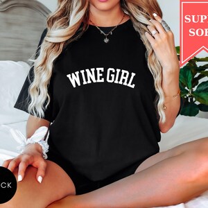Pode incluir: Camiseta preta com o texto "WINE GIRL" em letras brancas. Uma faixa vermelha no canto superior direito diz "SUPER SOFT". Um c&iacute;rculo preto no canto inferior esquerdo diz "BLACK".