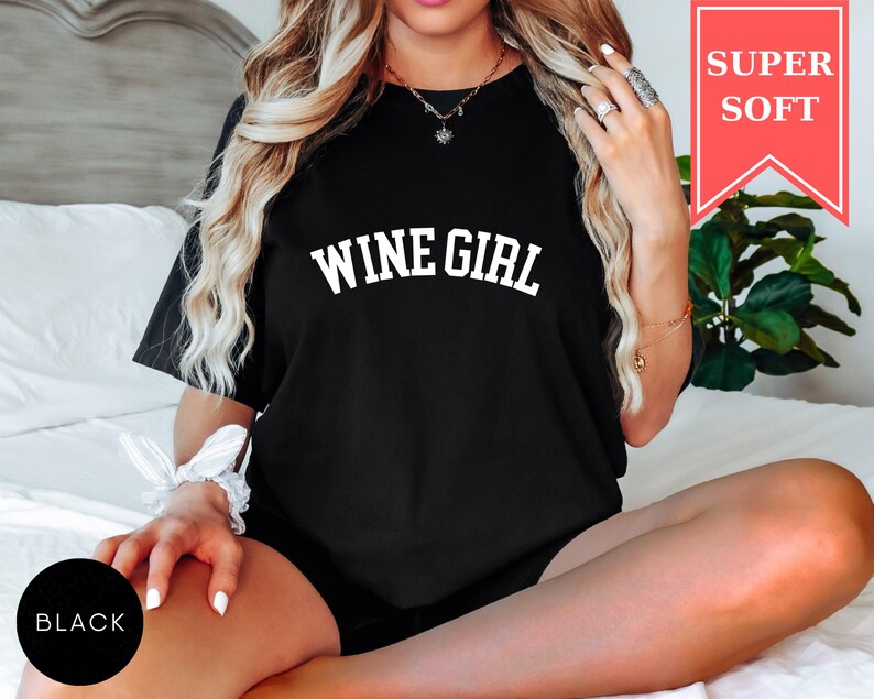 Pode incluir: Camiseta preta com o texto "WINE GIRL" em letras brancas. Uma faixa vermelha no canto superior direito diz "SUPER SOFT". Um c&iacute;rculo preto no canto inferior esquerdo diz "BLACK".