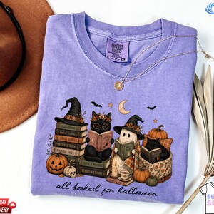 A purple t-shirt with a Halloween-themed illustration of cats dressed as witches and wizards, surrounded by books, pumpkins, and bats, along with the text "all hooked for Halloween".