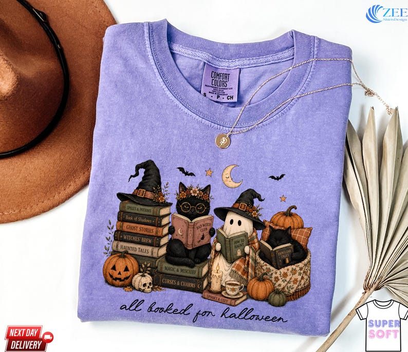 A purple t-shirt with a Halloween-themed illustration of cats dressed as witches and wizards, surrounded by books, pumpkins, and bats, along with the text "all hooked for Halloween".