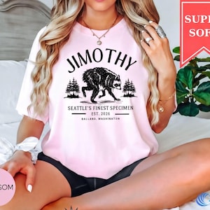 A woman wearing a pink t-shirt with a graphic design of a bear and text that says "JimOTHY Seattle's Finest Specimen EST. 2016 BALLARD, WASHINGTON".