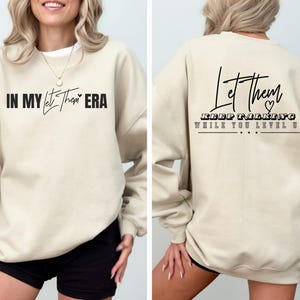 May include: A beige crewneck sweatshirt featuring black text. The front displays the phrase IN MY Let Them ERA. The back features the text Let Them KEEP TALKING WHILE YOU LEVEL UP with a small heart icon and three dots below the lettering.