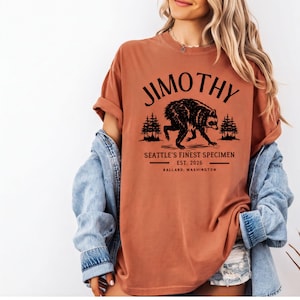 A woman wearing a red t-shirt with a graphic design of a bear and text that says "Jimothy, Seattle's Finest Specimen, EST. 2026, BALLARD, WASHINGTON".