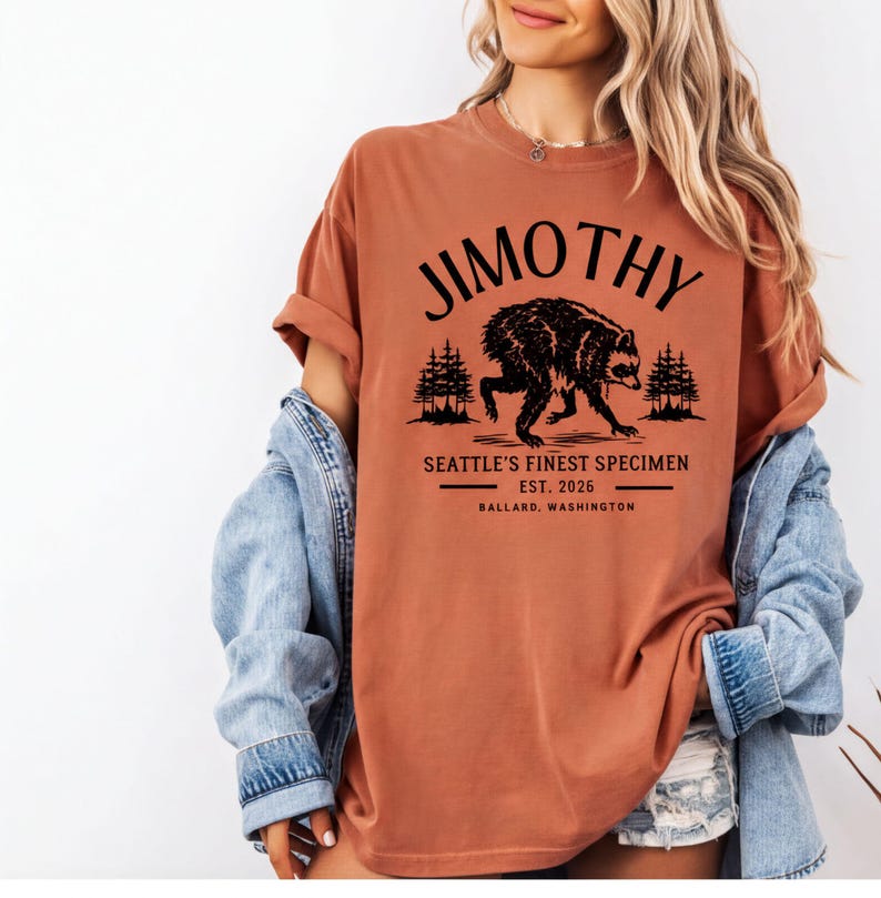 A woman wearing a red t-shirt with a graphic design of a bear and text that says "Jimothy, Seattle's Finest Specimen, EST. 2026, BALLARD, WASHINGTON".