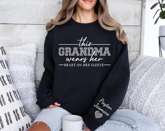 May include: A black long-sleeve sweatshirt featuring white text that reads this GRANDMA wears her HEART ON HER SLEEVE. The word GRANDMA and a heart icon on the left sleeve are filled with a white and black leopard print pattern. The sleeve also displays the names Payton and Jasmine in white script.