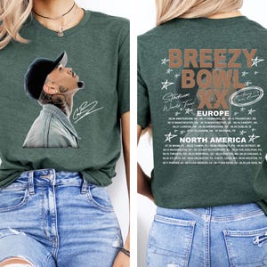 May include: A dark green t-shirt featuring a portrait of a person wearing a baseball cap and denim jacket. The back of the shirt displays the text "BREEZY BOWL XX Stadium World Tour" with a list of European and North American tour dates.