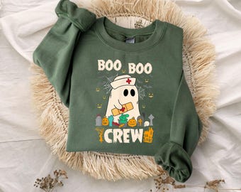 May include: An olive green crewneck sweatshirt featuring a graphic of a ghost wearing a nurse cap with a red cross, holding a bandage. The text reads BOO BOO CREW. The design includes small pumpkins, tombstones, a skull, and spider webs.