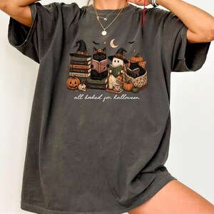 A person wearing a gray t-shirt with a Halloween-themed graphic design featuring cats, pumpkins, and a book.