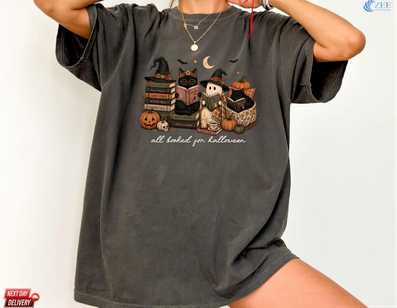 A person wearing a gray t-shirt with a Halloween-themed graphic design featuring cats, pumpkins, and a book.
