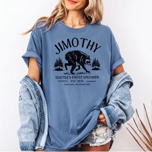 A woman wearing a blue t-shirt with a graphic design of a bear and text that says "Jimothy, Seattle's Finest Specimen, Est. 2006, Ballast, Washington".