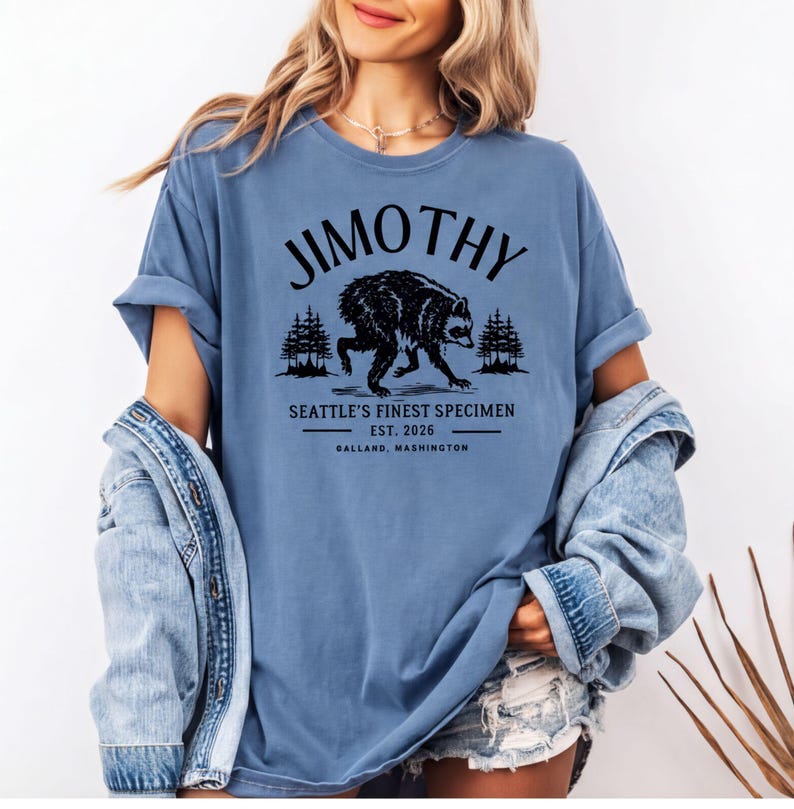 A woman wearing a blue t-shirt with a graphic design of a bear and text that says "Jimothy, Seattle's Finest Specimen, Est. 2006, Ballast, Washington".